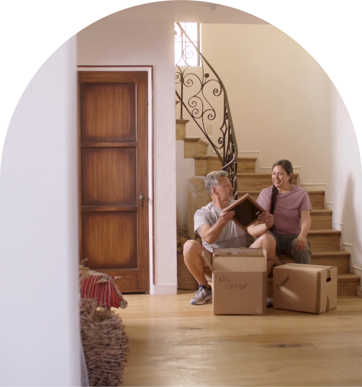  A man and a woman sitting on a staircase amidst moving boxes in their new home. The man holds a picture, and the woman, with a joyful expression, gazes at the image, symbolizing fond memories in their new space.