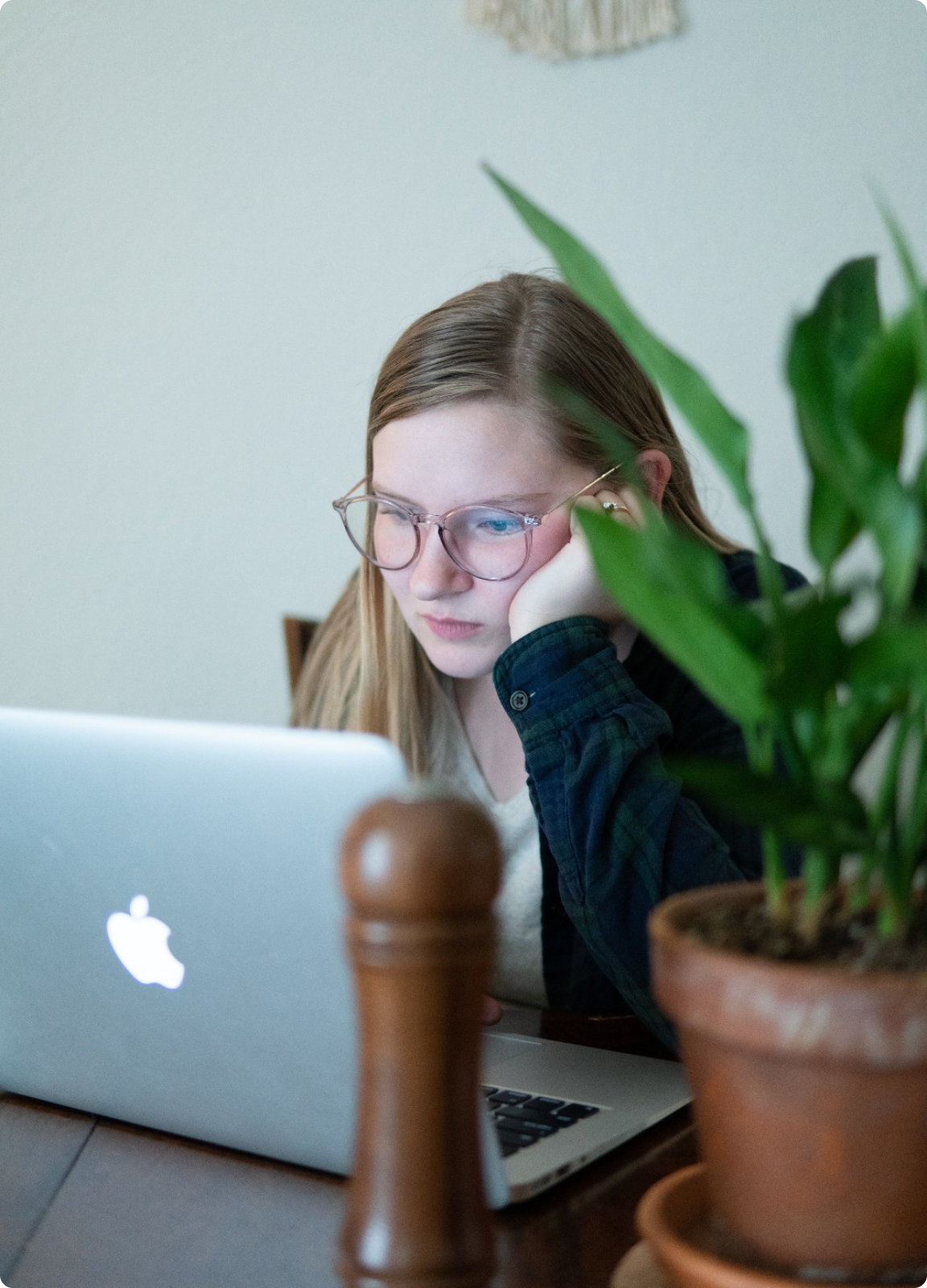 A focused woman with glasses, her elbow resting on the table, and her hand supporting her chin as she concentrates on her laptop. Her expression reflects a sense of seriousness and concentration.