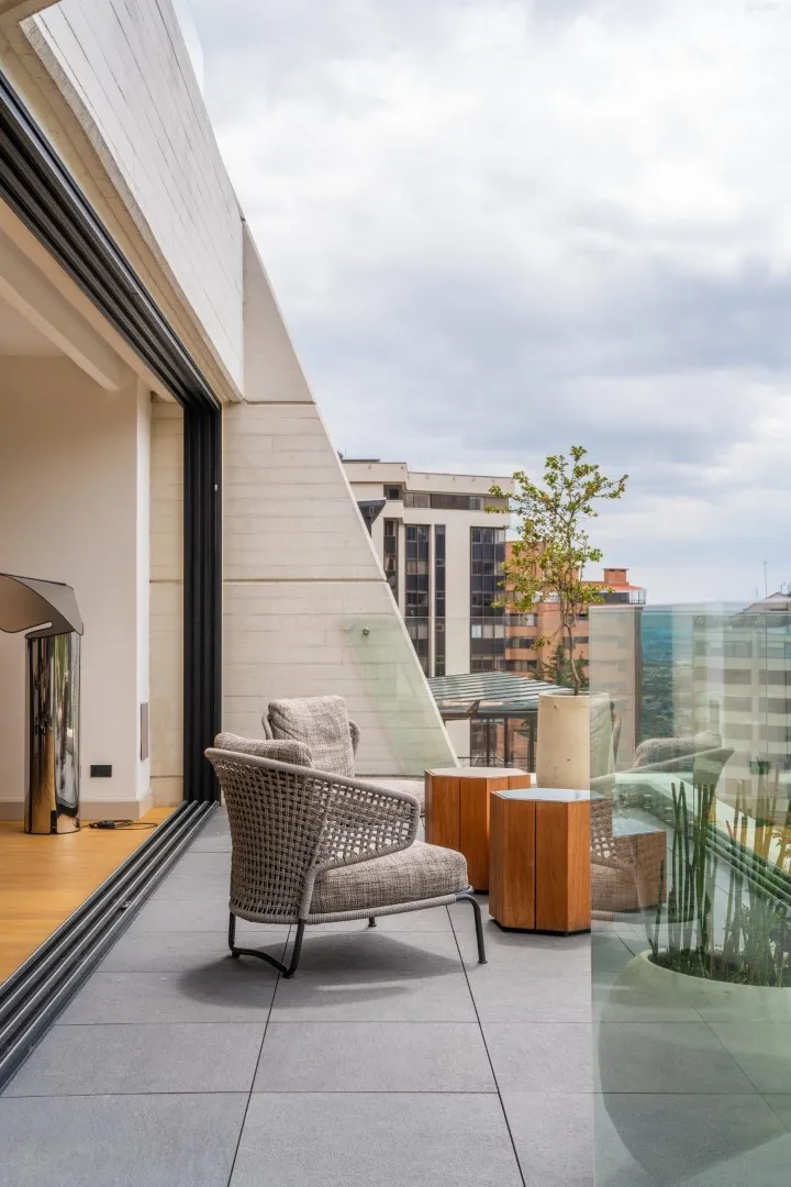 Balcón moderno con silla de mimbre, mesas de madera y plantas en macetas con vista a edificios urbanos.