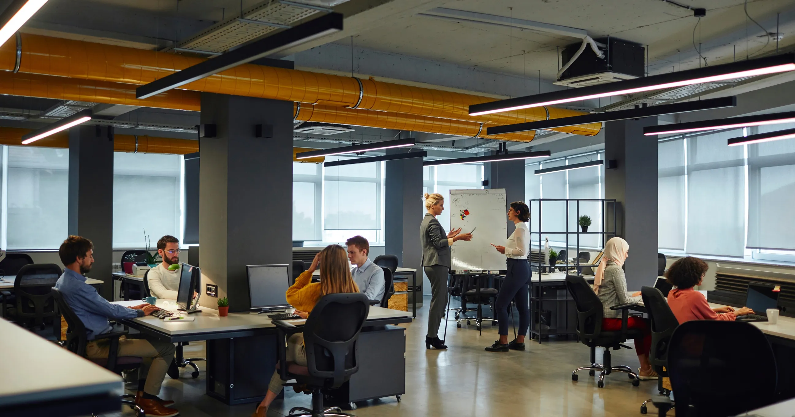 Espaço de escritório moderno com pessoas trabalhando e duas mulheres conversando perto de um quadro branco.