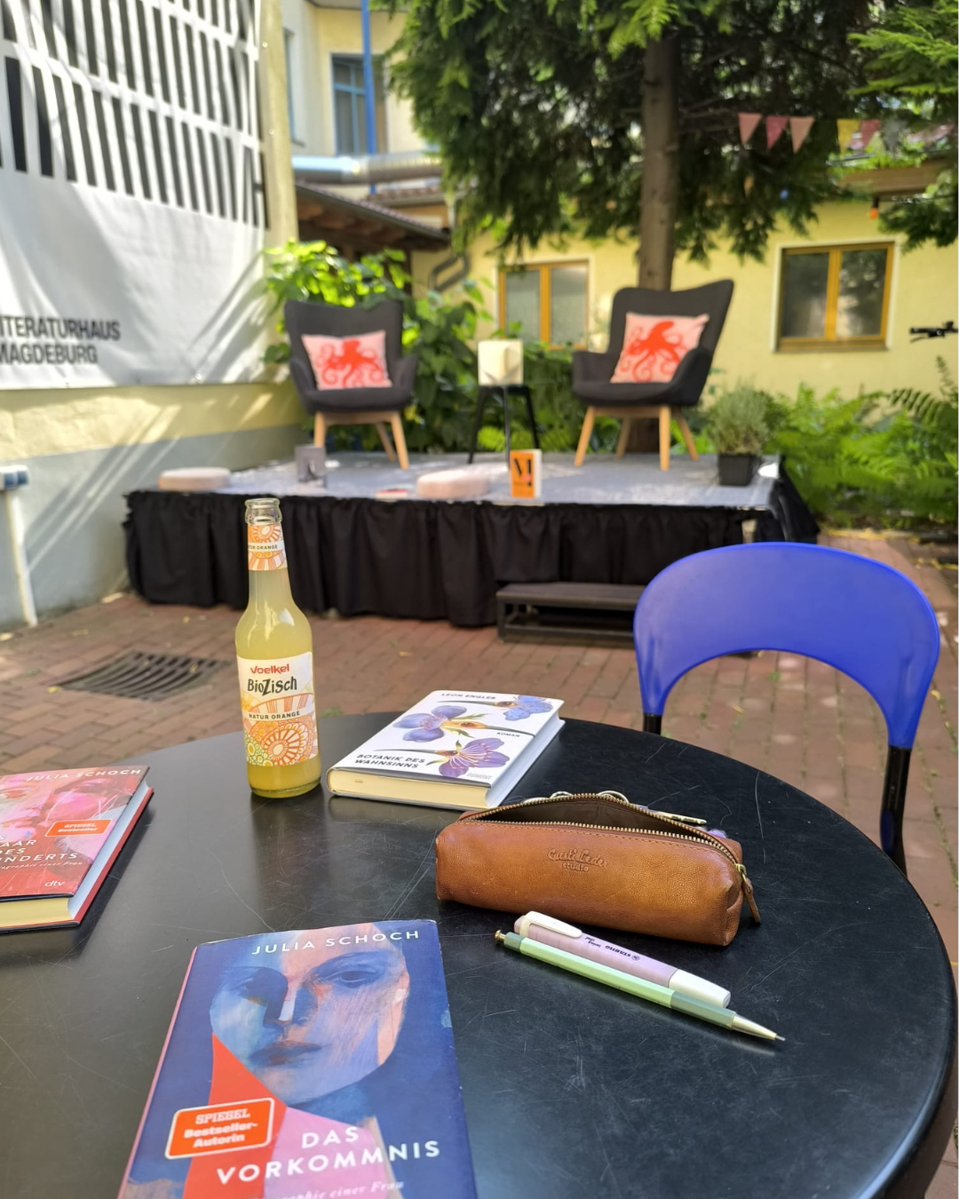 Eine Bühne mit zwei Lesesesseln im Hintergrund. Im Vordergrund ein runder Tisch mit Büchern, Stiften und Limonade.