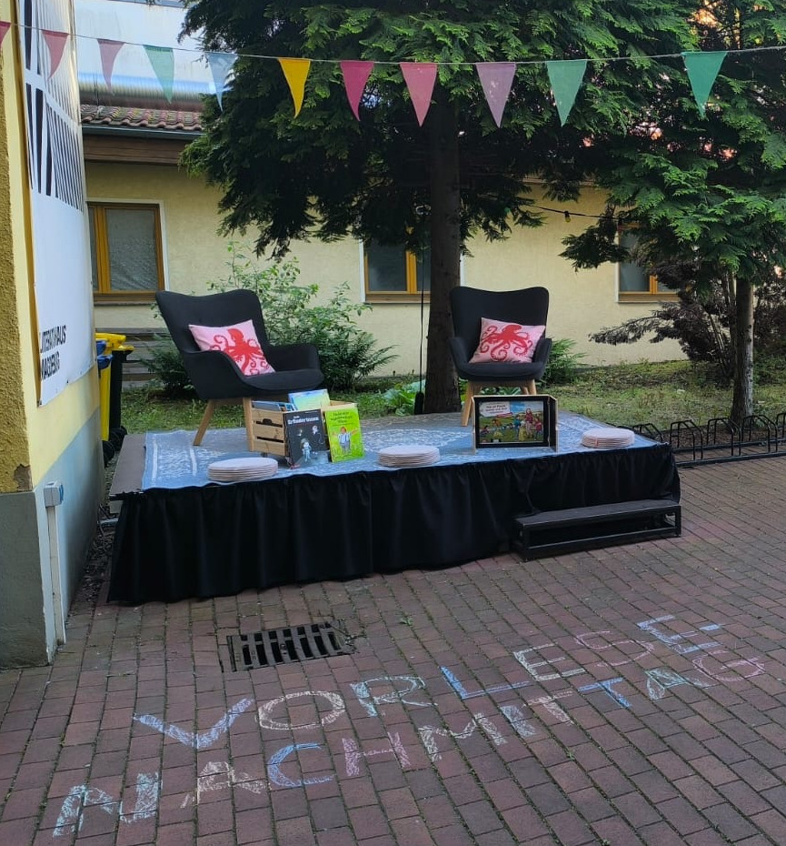 Unsere kleine Bühne im Hinterhaus mit zwei Sesseln, bunter Wimpelkette und dem Kreideschriftzug "Vorlese Nachmittag" auf dem Boden.