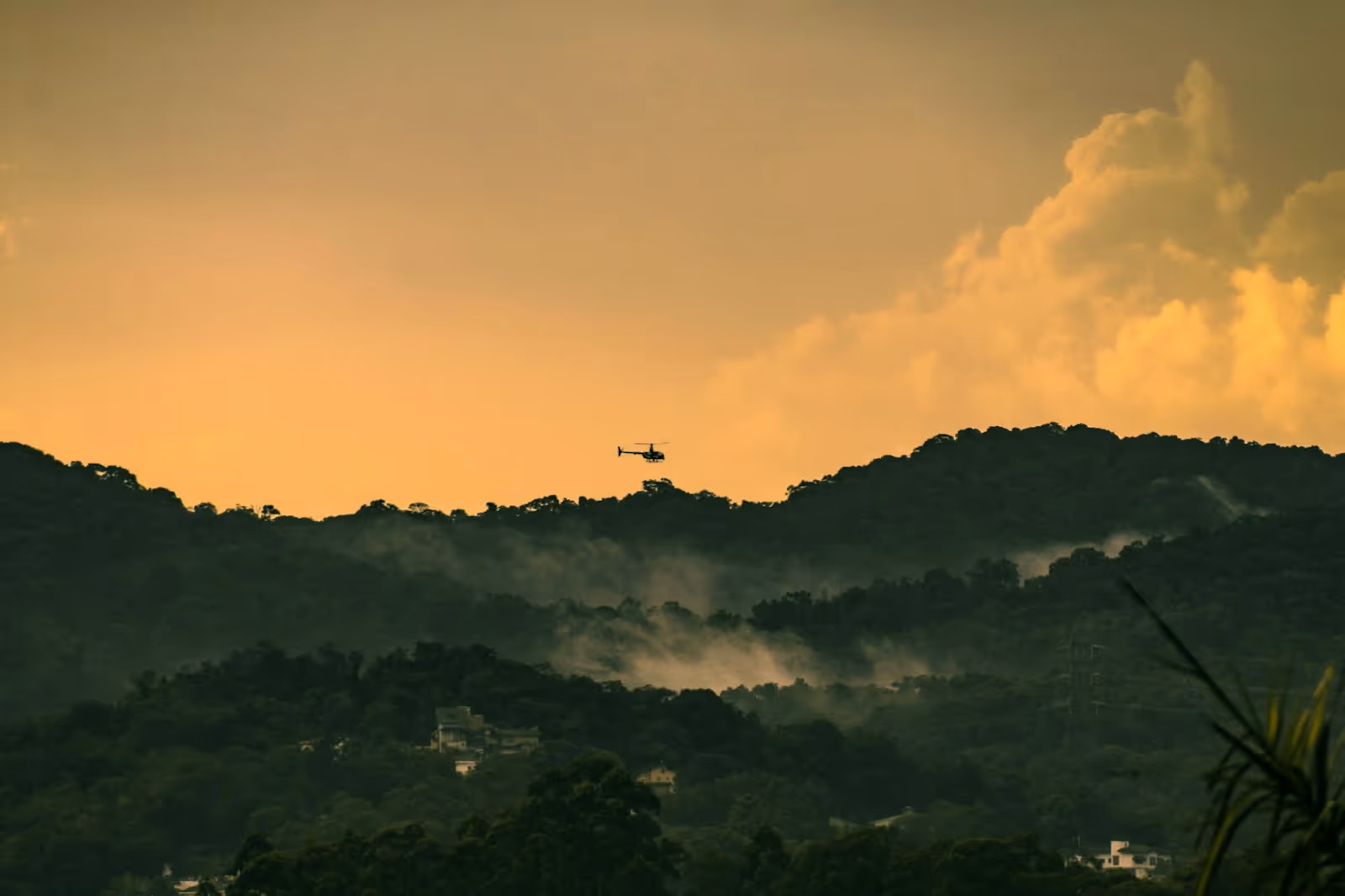 silhouette of a helicopter flying over forested mountains at dusk
