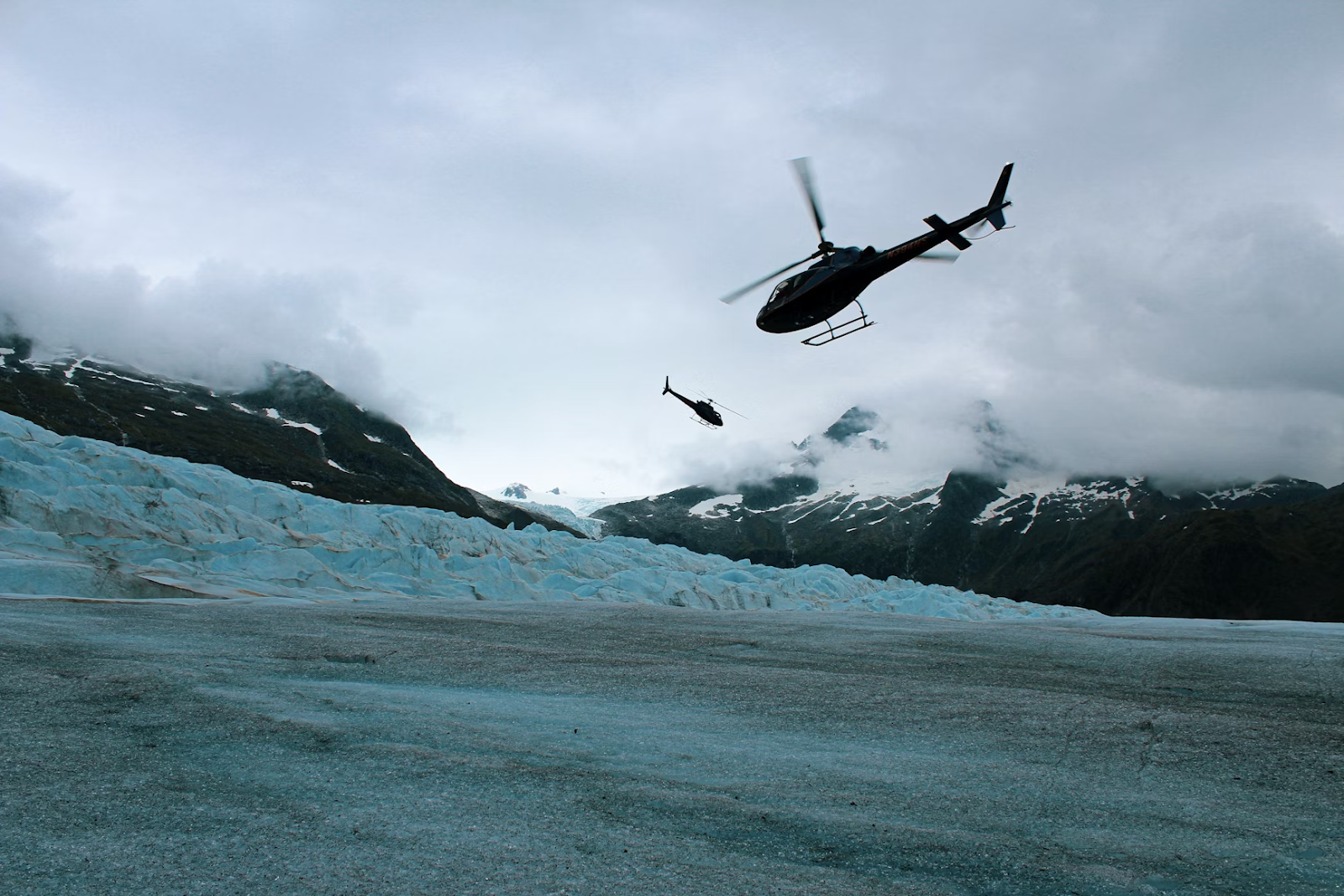 two black helicopters flying over ice-covered mountains on a cloudy day
