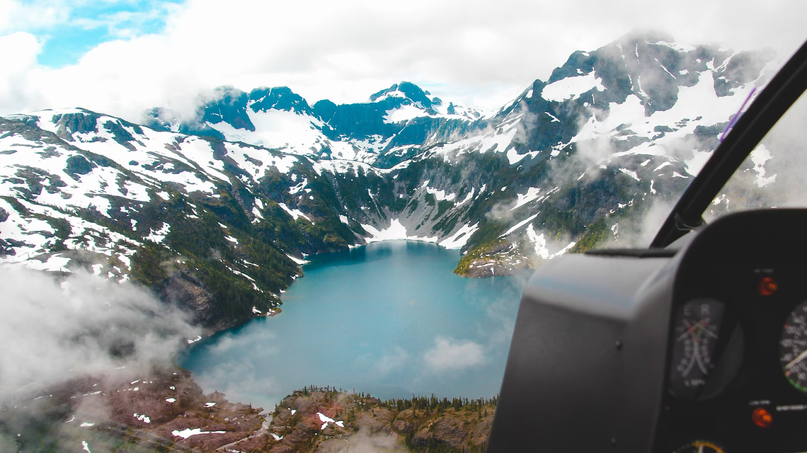 view of snowy mountains and a glacial lake from inside a helicopter cockpit