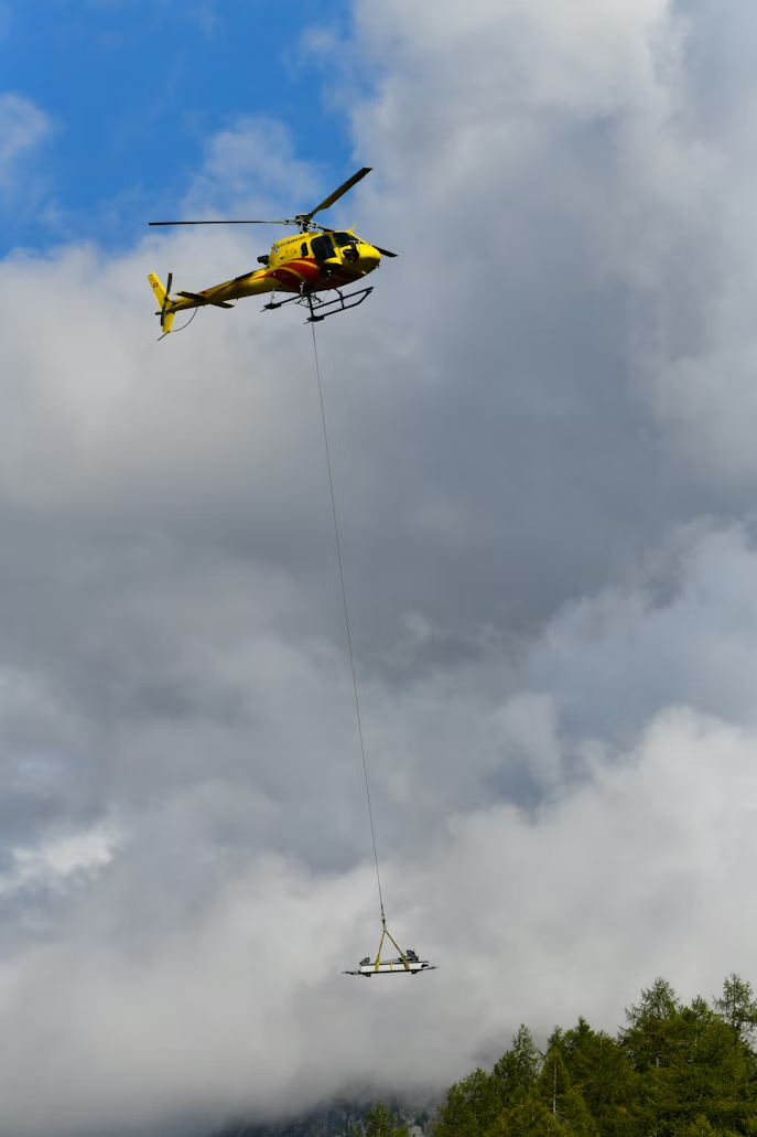 yellow and red utility helicopter carrying cargo on a long line above a forest on a cloudy day