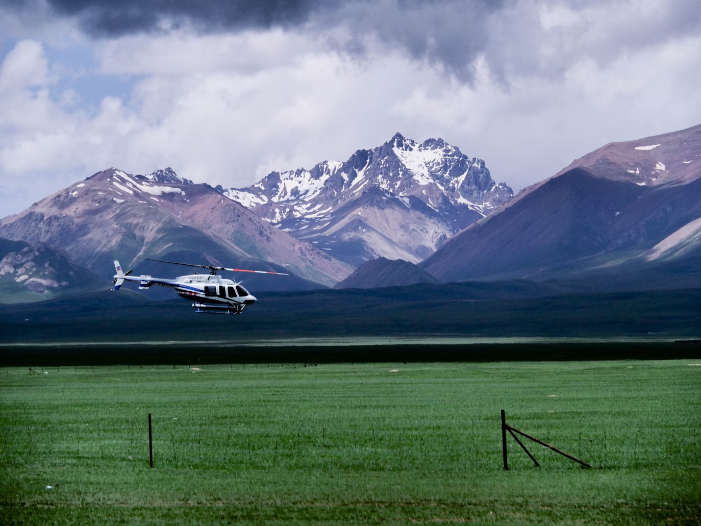 white helicopter flying low over a grassy field with snowy mountains in the background