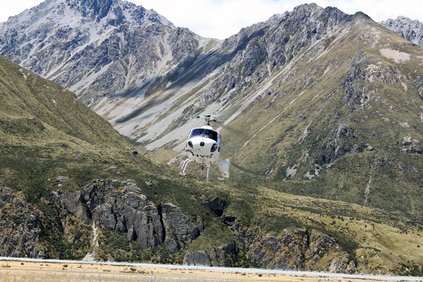 white helicopter flying low over the ground in front of grassy mountains in New Zealand