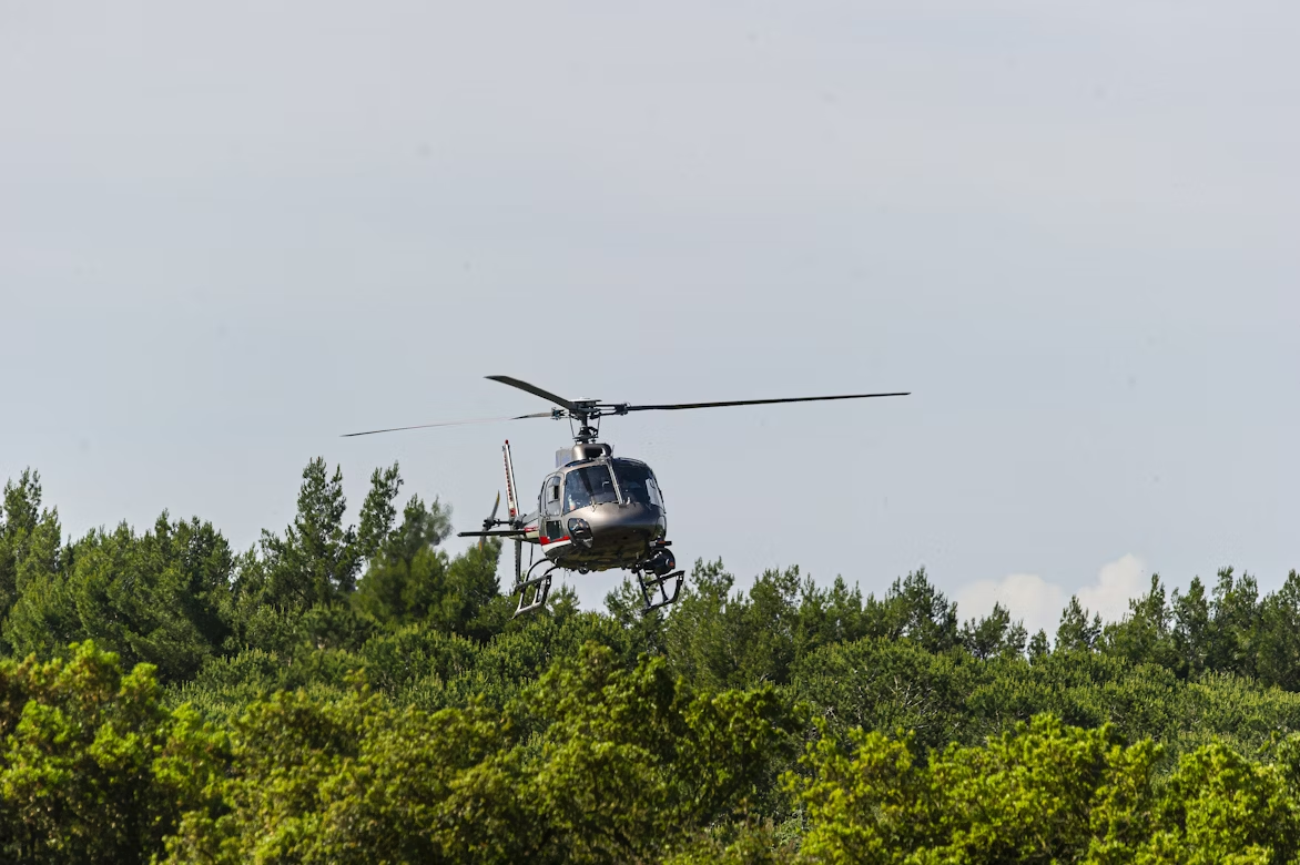 red and gray helicopter flying low over a green forest on a gray sky day