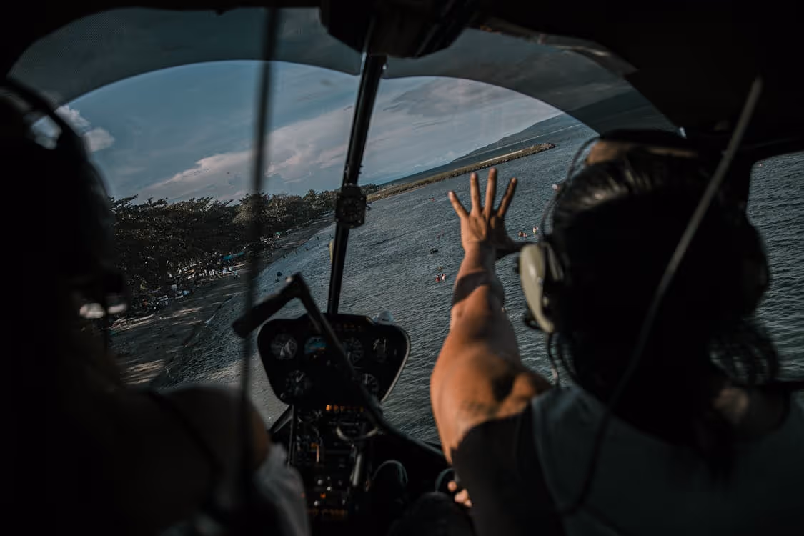 view from inside a helicopter cockpit of a pilot with an outstretched arm flying low over a beach