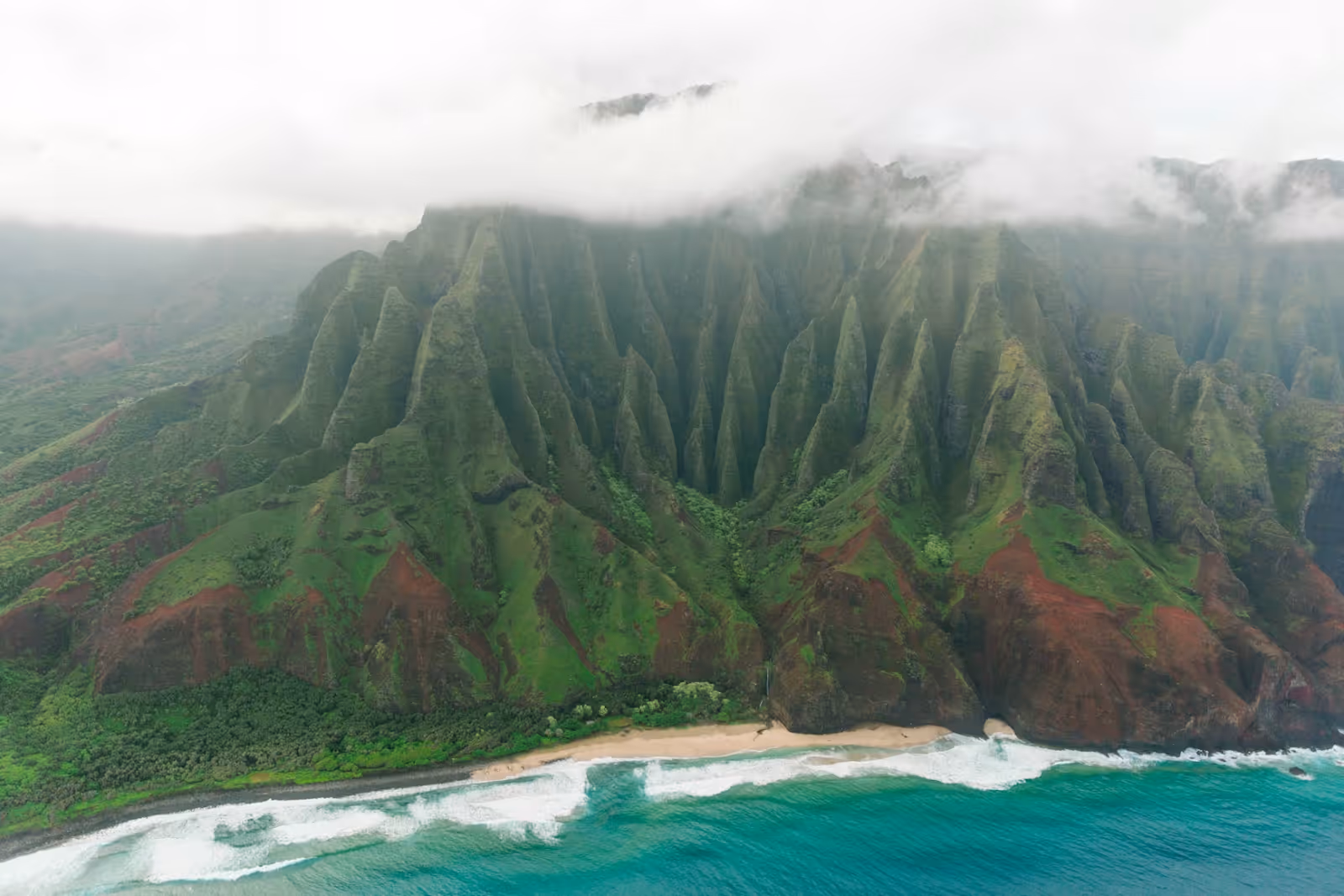 aerial view of a small beach surrounded by green and brown mountains extending into the clouds