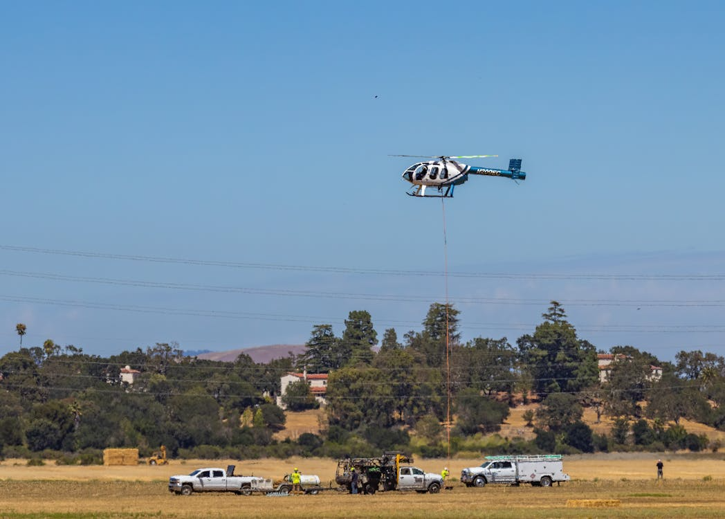 white and blue helicopter hovering over a power line with utility workers and trucks below