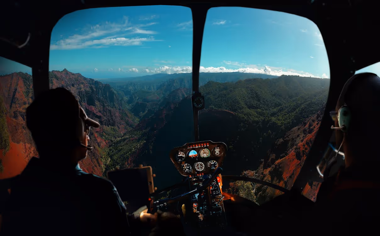 two pilots inside a helicopter with large windows flying over grassy mountains