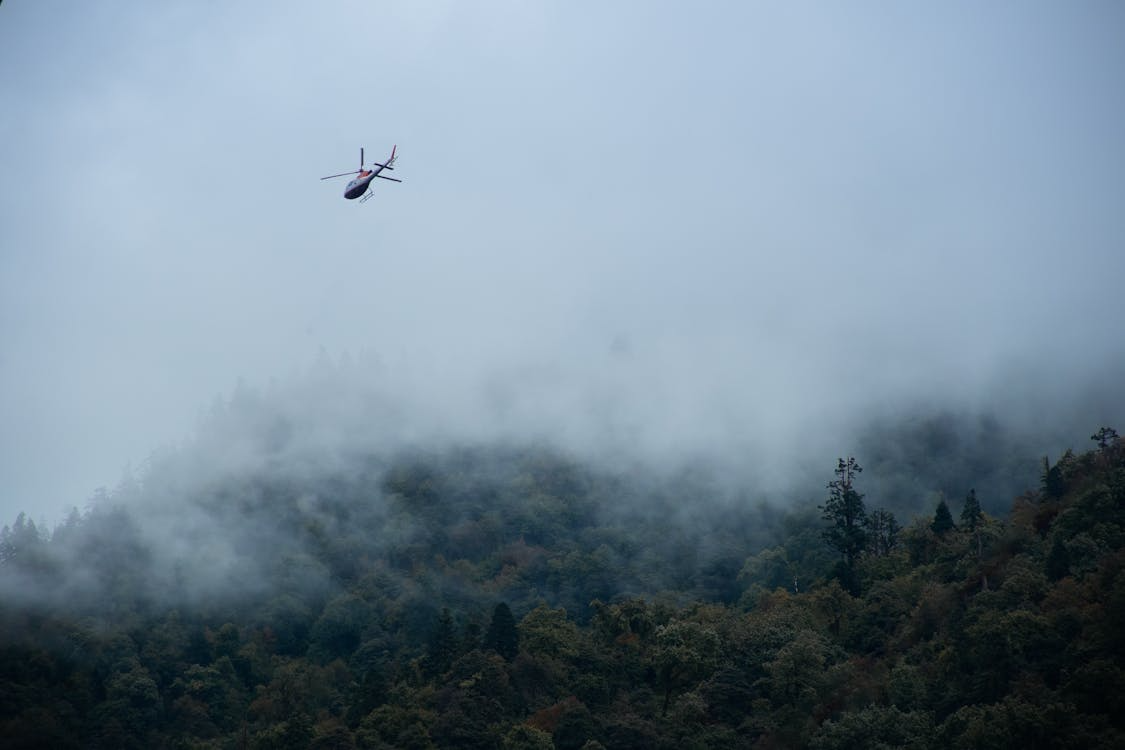 red and white helicopter flying over foggy tree-covered mountains