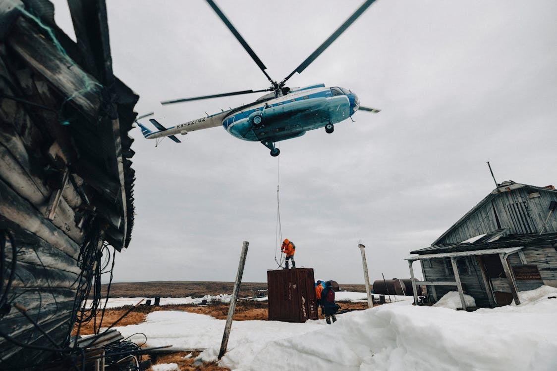 blue and white helicopter hovering while three men disconnect its cargo in a remote winter landscape