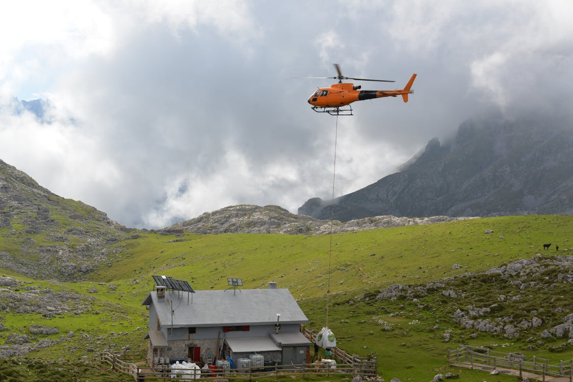 orange helicopter using a long line to deliver cargo to a remote house in the mountains of Europe