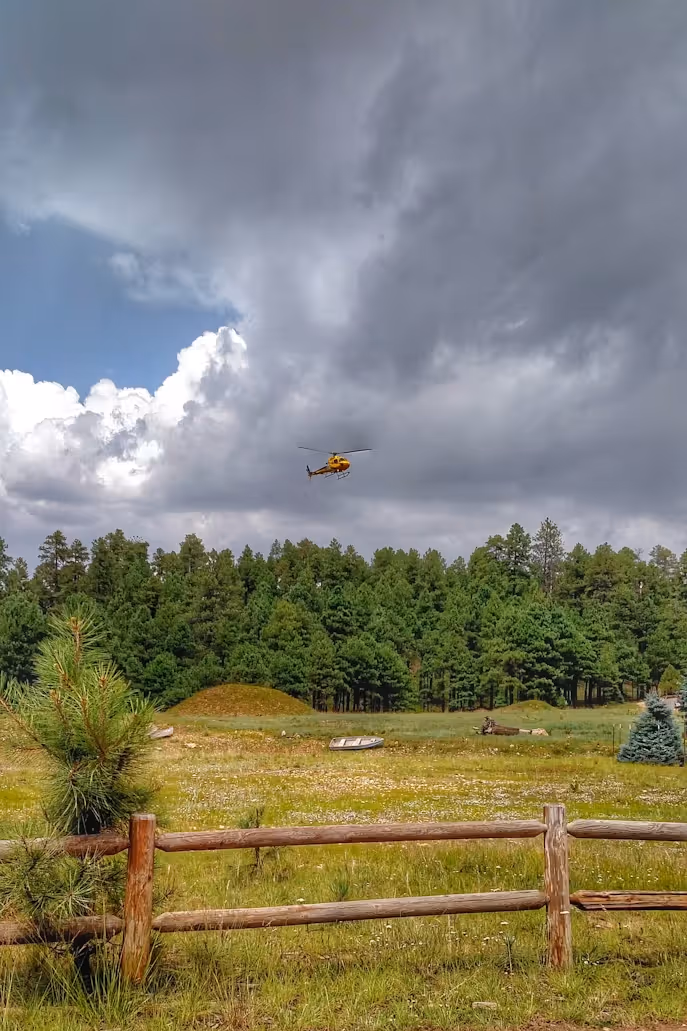 red and yellow helicopter flying over a forest and pasture under dark clouds