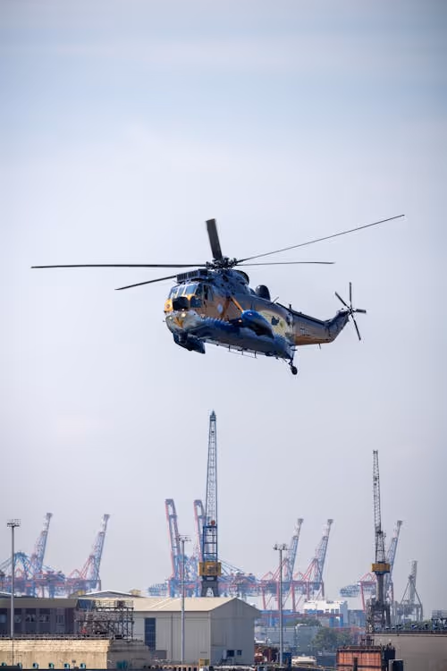 blue and yellow helicopter flying over a harbor with ground cranes in the background