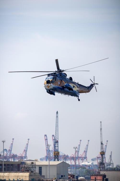 blue and yellow helicopter flying over a harbor with ground cranes in the background