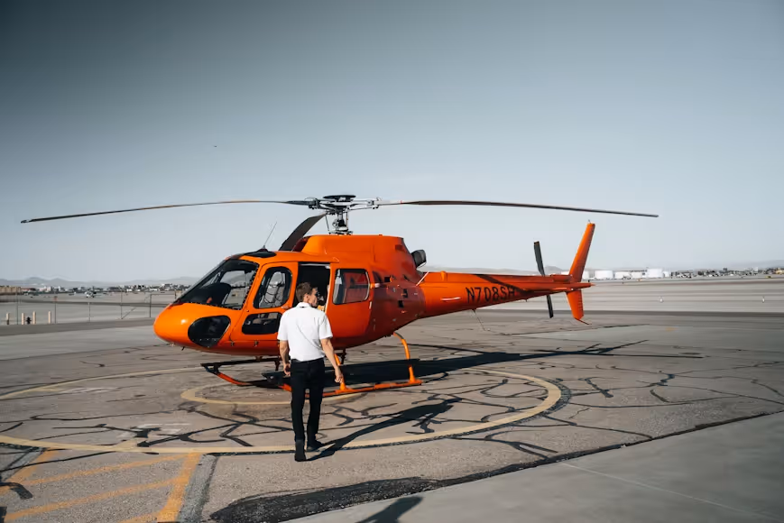 male pilot walking toward an orange helicopter resting on a concrete helipad