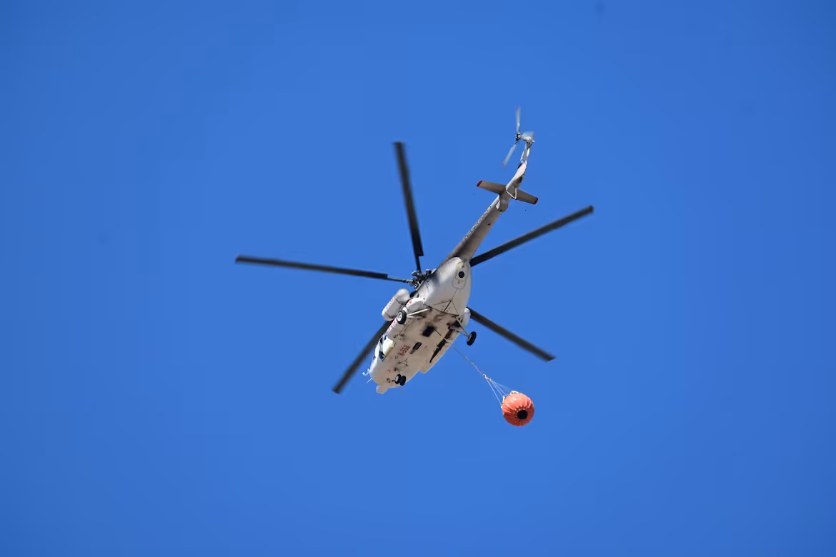white helicopter with black blades carrying a Bambi Bucket under blue skies