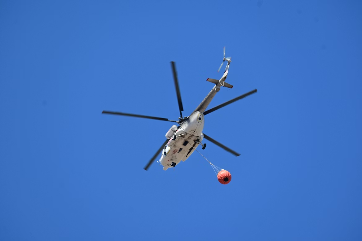 white helicopter with black blades carrying a Bambi Bucket under blue skies