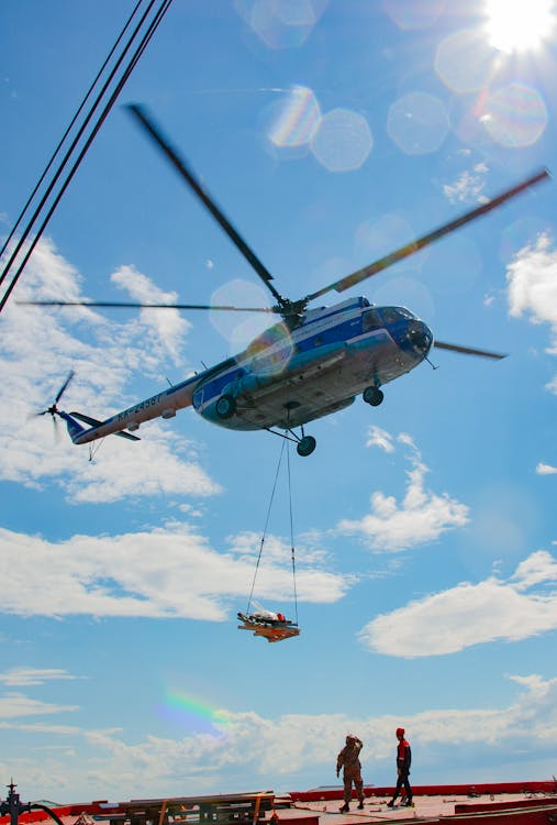 blue and white helicopter lowering cargo on ropes onto a rooftop with ground crews looking on