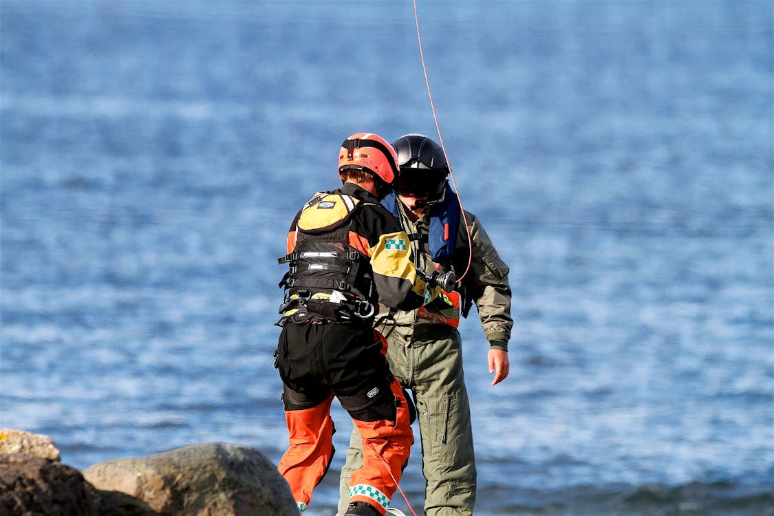 emergency response worker attaching a helicopter line to a rescued individual as they stand in front of water