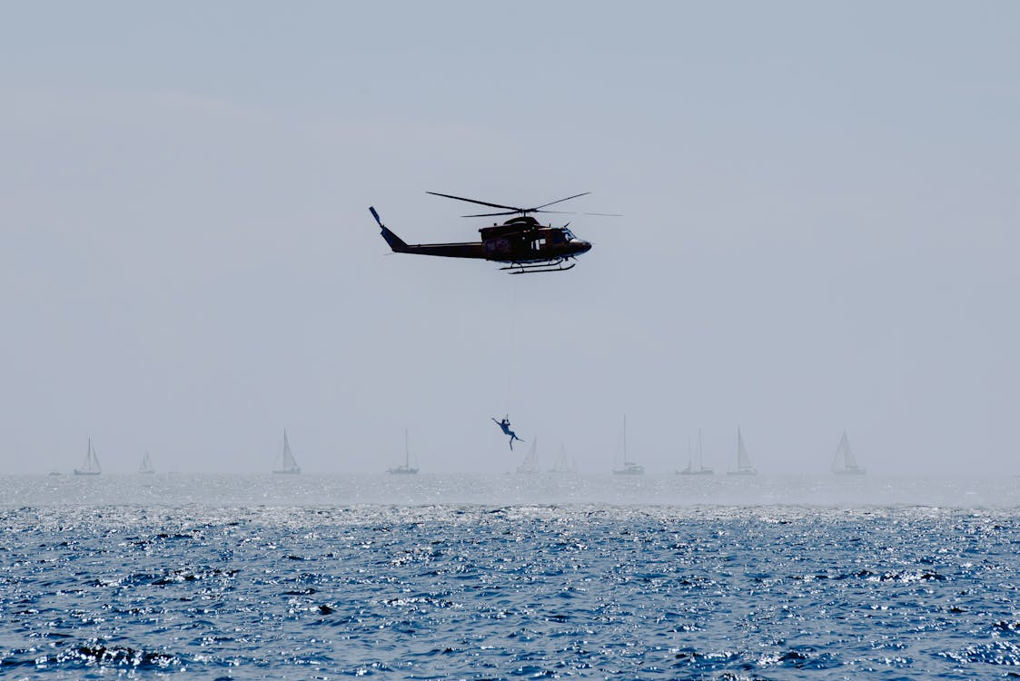 rescuer hanging over choppy water on a long line as a black helicopter hovers above