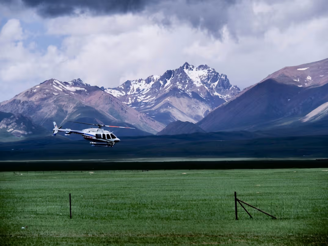 white, blue, and red helicopter landing in a grassy field in front of snow-covered mountains on a cloudy day