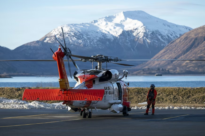 Coast Guard helicopter and crew member on a tarmac in front of water and a snow-covered mountain