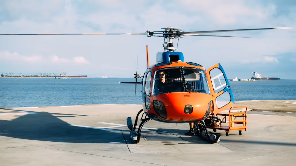 orange helicopter resting on a concrete tarmac by the ocean