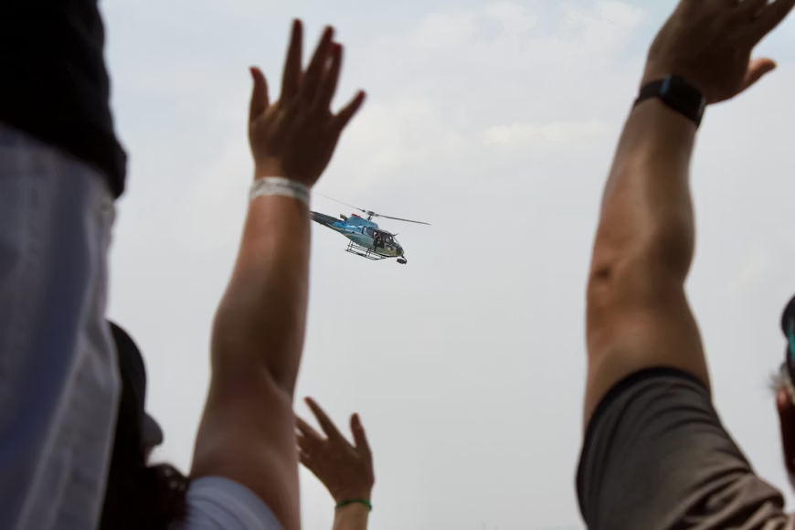 crowd raising their arms to wave at a blue helicopter flying overhead on a cloudy day