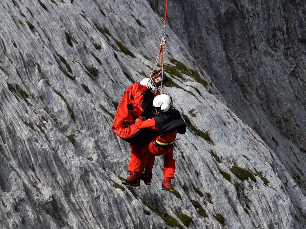 rescuers suspended from a helicopter hoist rescuing a person from the side of a mountain