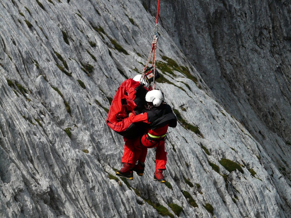 rescuers suspended from a helicopter hoist rescuing a person from the side of a mountain