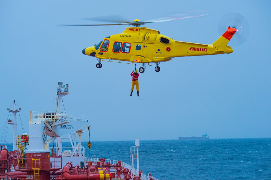 yellow rescue helicopter hovers above a ship while a hoist lifts a person from the ship deck