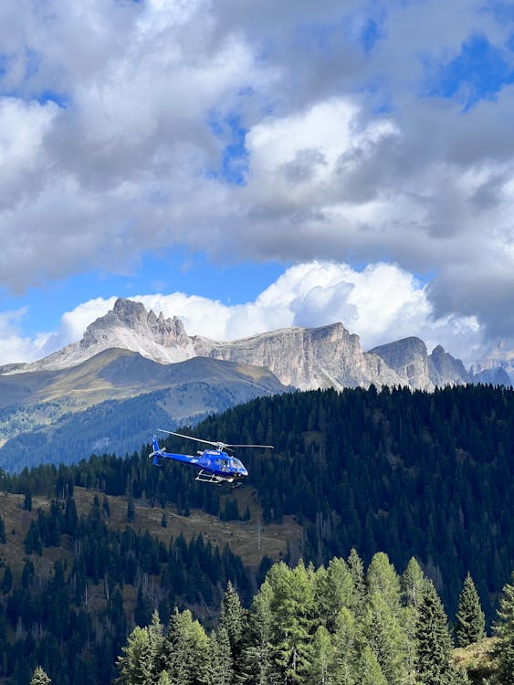 blue helicopter flying low over a tree line with mountains in the distance and clouds overhead