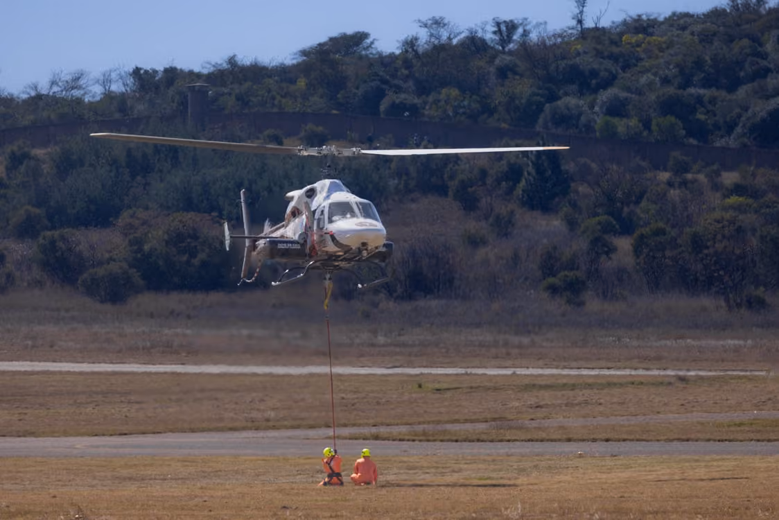 white helicopter hovering above a grassy field with two crew members on the ground in orange safety gear