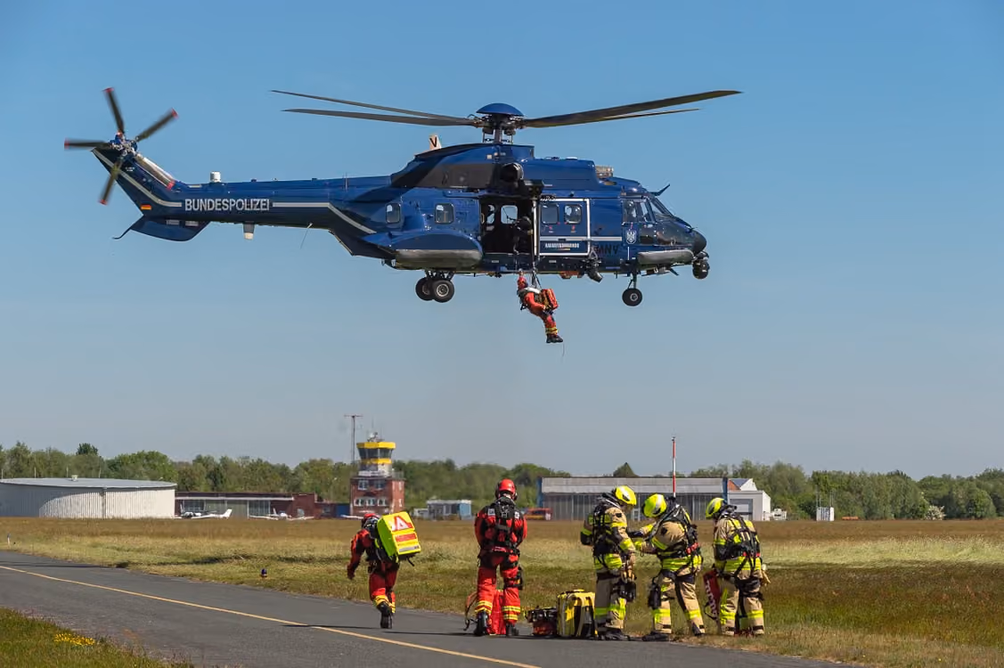 police helicopter conducting hoist training with rescue personnel in an outdoor field setting under blue skies