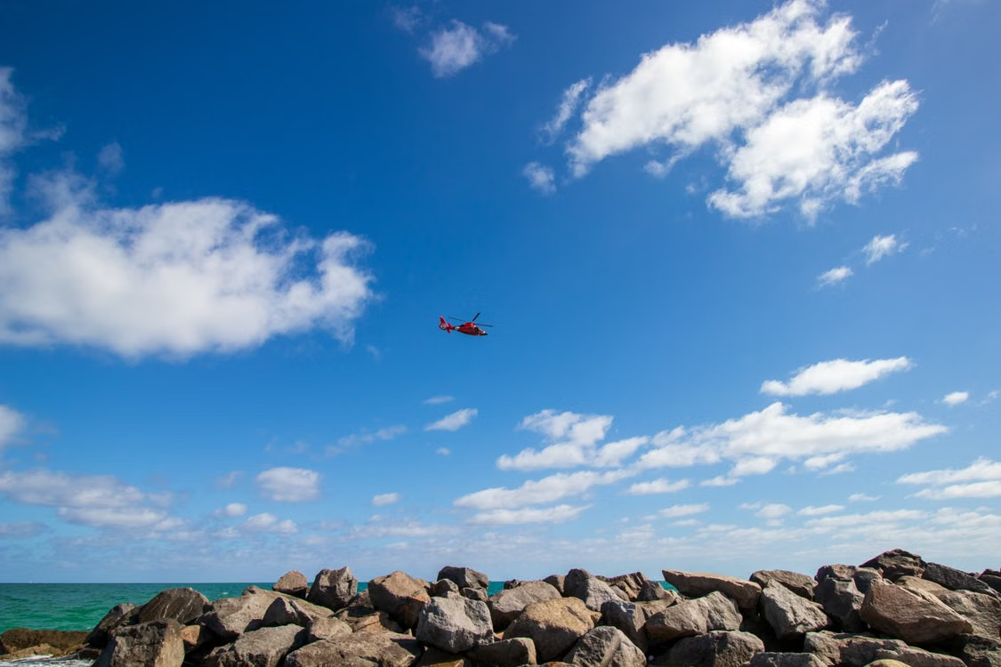 red helicopter flying over a rocky Miami coastline under a bright blue sky