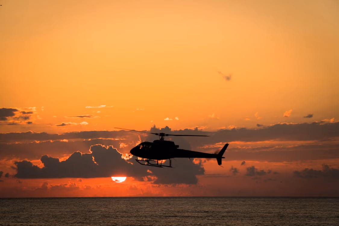 dark silhouette of a helicopter flying low over the ocean against an orange cloudy sunset
