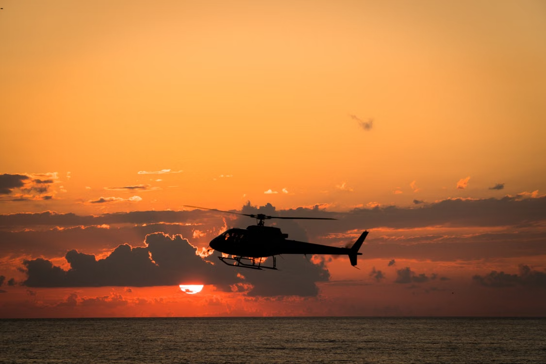 dark silhouette of a helicopter flying low over the ocean against an orange cloudy sunset