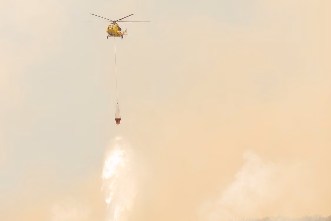 yellow firefighting helicopter dropping water from a Bambi Bucket to combat wildfires amidst smoky skies