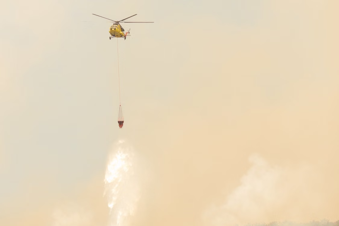 yellow firefighting helicopter dropping water from a Bambi Bucket to combat wildfires amidst smoky skies
