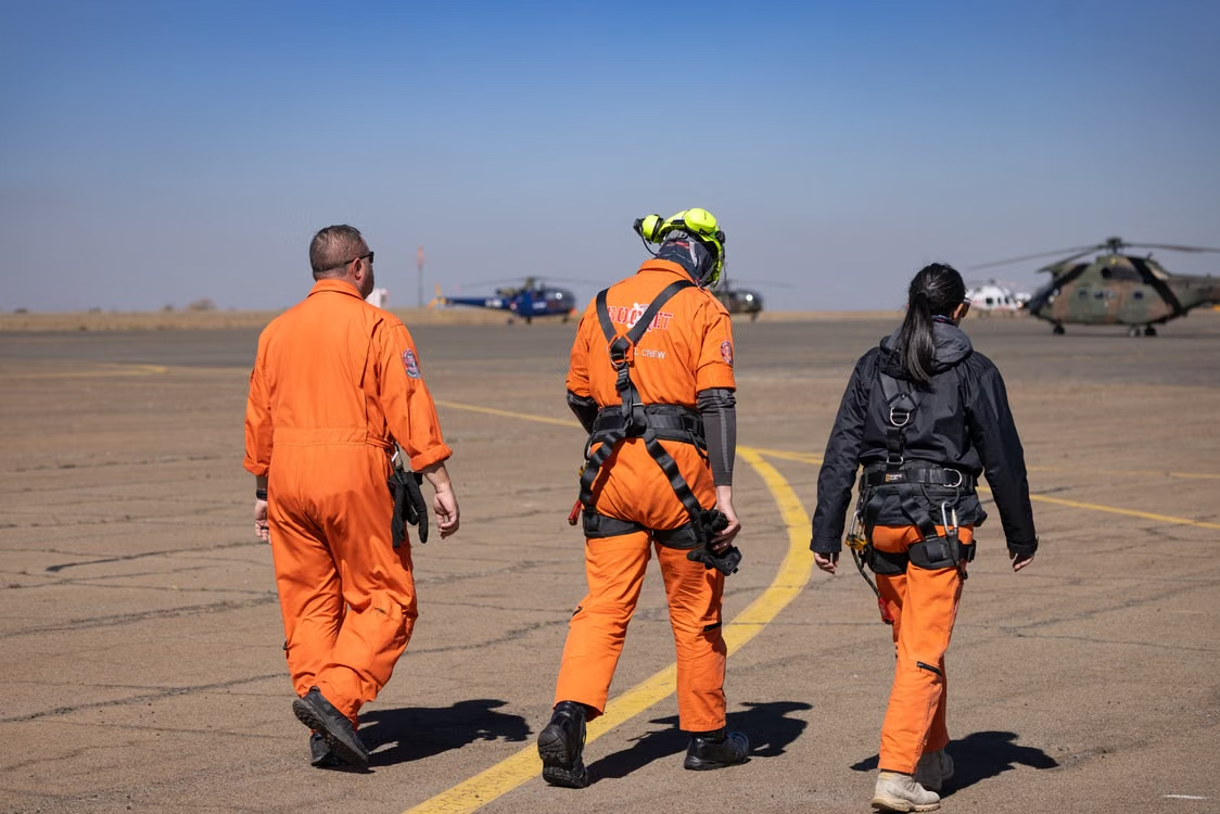three rescue personnel in orange uniforms walking on a tarmac toward helicopters under blue skies