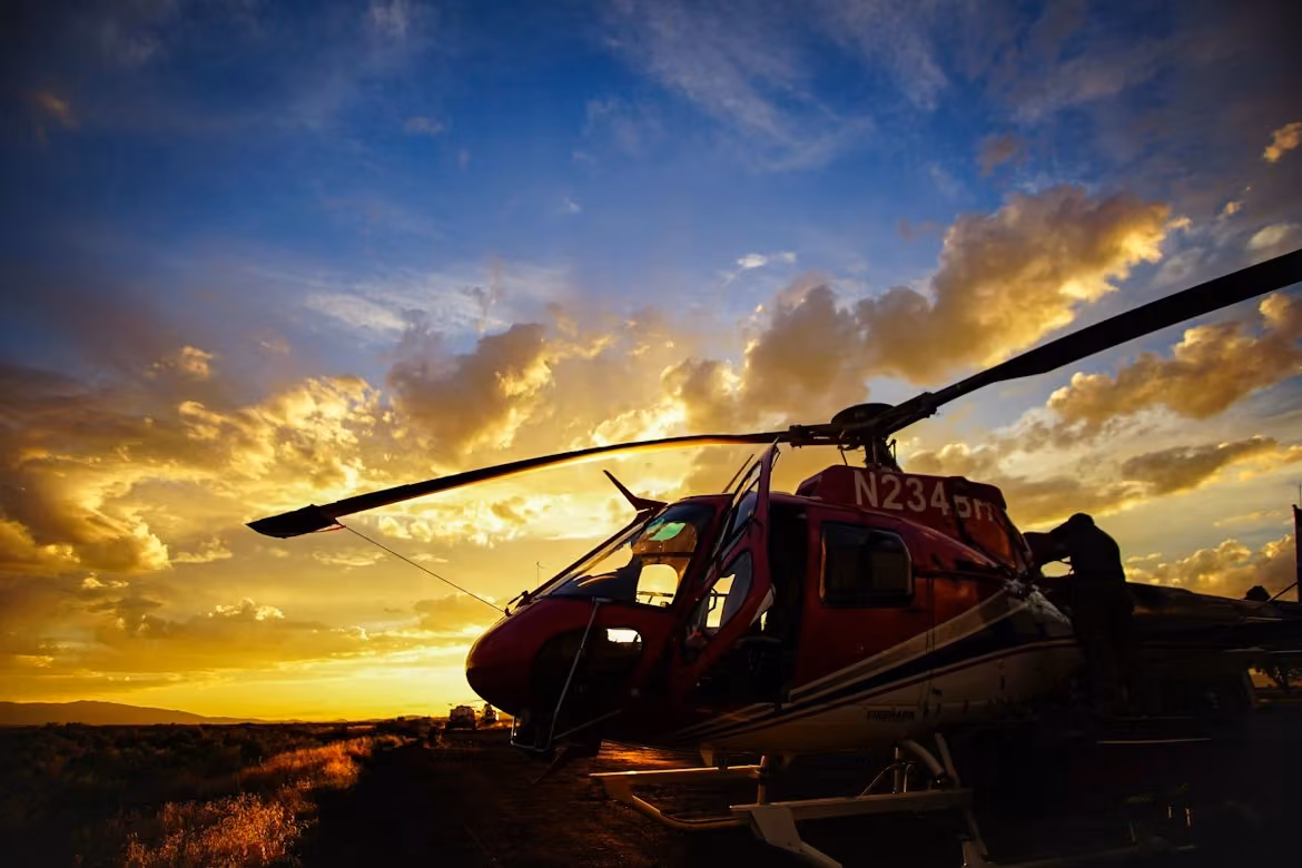 red and white helicopter parked in a remote landscape at sunset as a crew member works on the aircraft