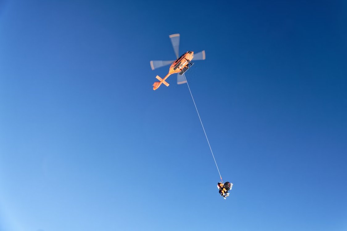 orange emergency helicopter performing a rescue operation with a rescuer suspended in mid-air against a clear blue sky