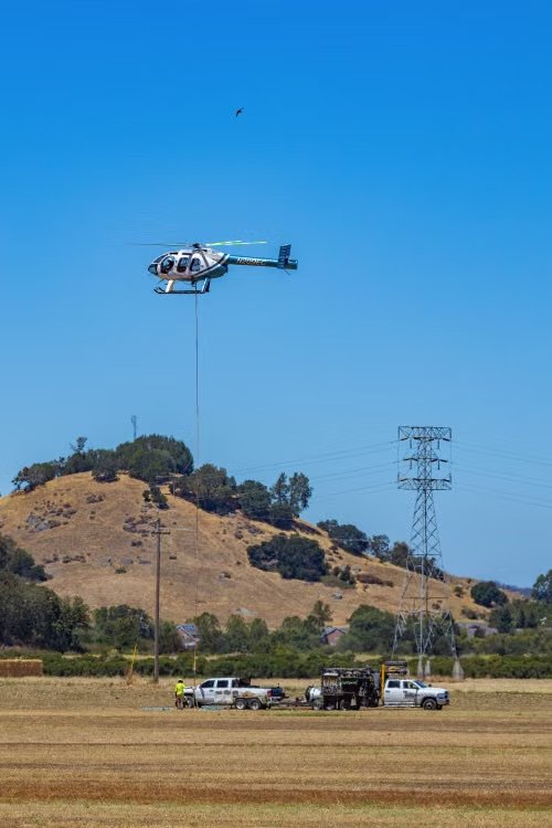 white and blue utility helicopter hovering over powerlines and utility workers in a rural landscape under a clear blue sky