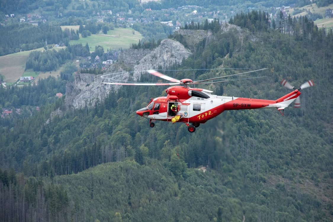 red and white rescue helicopter flying over the scenic Tatra Mountains in Poland with an open door as a responder prepares for a rescue mission