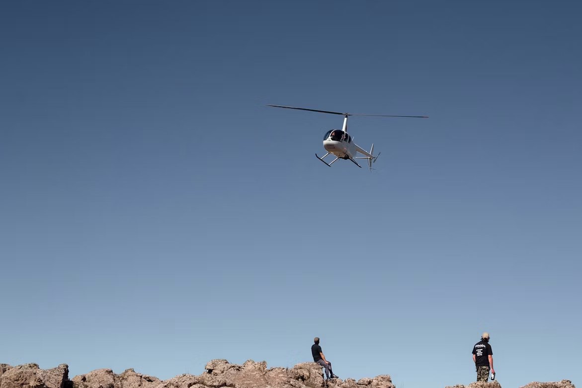 white utility helicopter flying over two men standing and sitting on large rocks under clear blue skies